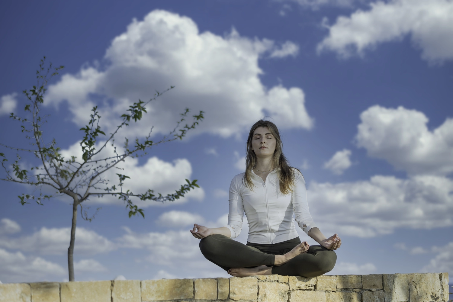 Yoga für Dich - Yoga Kurse - Eine Frau sitzt in einer meditativen Yoga-Pose auf einer niedrigen Backsteinmauer im Freien unter blauem Himmel mit weißen Wolken.